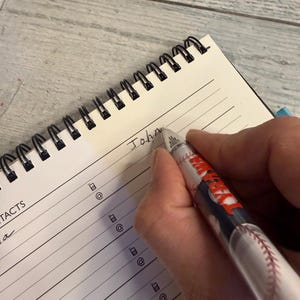 May include: A spiral-bound notebook with lined pages featuring a contacts section with phone and email icons. A pen with a baseball graphic and the word "TARAWA" printed on the barrel is writing the name "John" on the top line of the notebook.