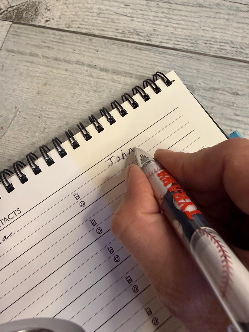 May include: A spiral-bound notebook with lined pages featuring a contacts section with phone and email icons. A pen with a baseball graphic and the word "TARAWA" printed on the barrel is writing the name "John" on the top line of the notebook.