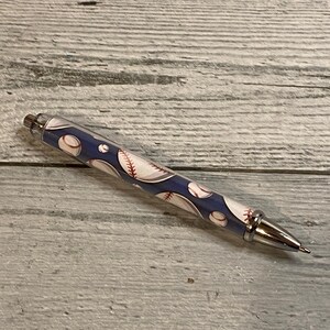 May include: A retractable ballpoint pen featuring a dark blue barrel decorated with a repeating pattern of white baseballs with red stitching. The pen includes a polished silver-tone metal tip, a matching silver-tone click mechanism, and a silver-tone clip.