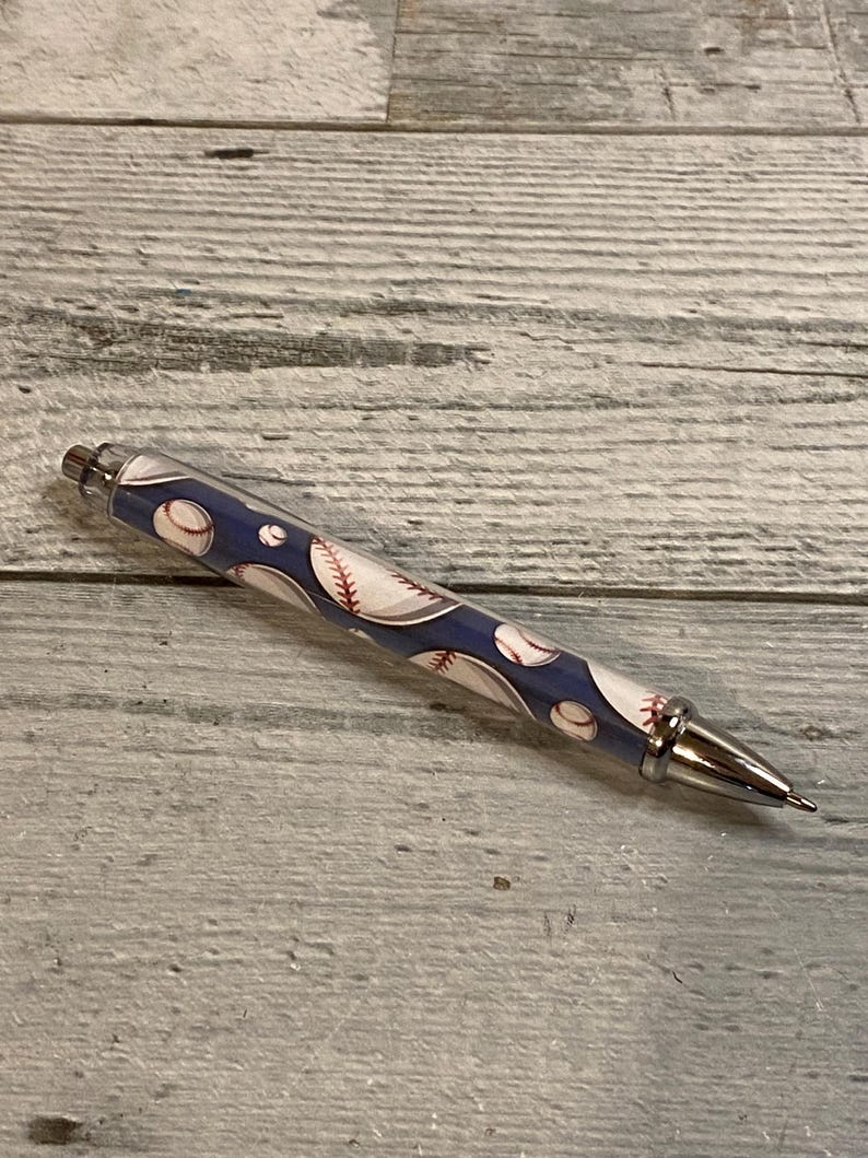 May include: A retractable ballpoint pen featuring a dark blue barrel decorated with a repeating pattern of white baseballs with red stitching. The pen includes a polished silver-tone metal tip, a matching silver-tone click mechanism, and a silver-tone clip.