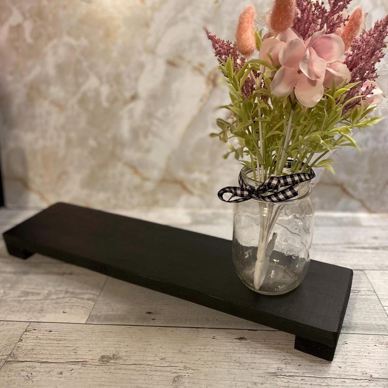 May include: A rectangular black wooden riser with short feet, paired with a clear glass mason jar containing a bouquet of pink and light purple artificial flowers, accented by a black and white checkered ribbon tied around the jar neck.