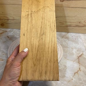 May include: A rectangular wooden board with a light brown finish and visible natural grain patterns. The surface is smooth and flat, showcasing the organic texture of the wood with darker streaks running vertically along the length of the piece.