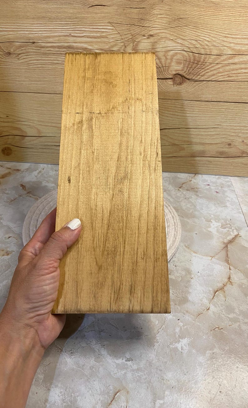 May include: A rectangular wooden board with a light brown finish and visible natural grain patterns. The surface is smooth and flat, showcasing the organic texture of the wood with darker streaks running vertically along the length of the piece.