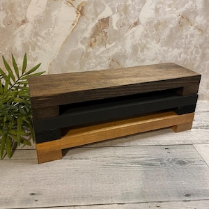 May include: A wooden riser with a dark brown top, black middle section, and natural wood base. The tiered design creates a visually appealing display for showcasing items. The wood grain is visible, adding to the rustic charm.