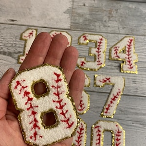 May include: A set of white felt baseball jersey numbers with red stitching and gold glitter trim. The numbers include 1, 3, 4, 6, 7, 8, and 0.