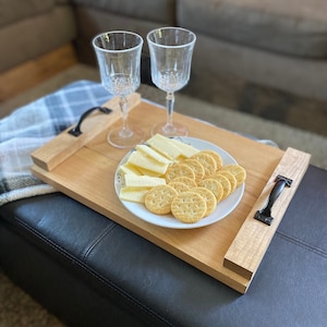 May include: A rectangular light-colored wooden serving tray with two black metal handles. On the tray sits a white plate containing round crackers and slices of yellow cheese, accompanied by two clear glass stemmed goblets for beverages.