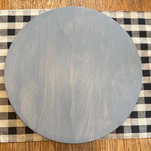 May include: A round, light blue wooden board. The board is set on a black and white checkered fabric. The wood grain is visible on the surface of the board. The board is a solid color with no other design elements.