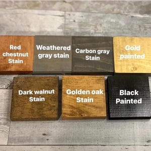 K&ouml;nnte beinhalten: Sieben quadratische Holzbl&ouml;cke in zwei Reihen, jeder mit einem eigenen Finish und wei&szlig;en Textbeschriftungen: Red chestnut Stain, Weathered gray stain, Carbon gray Stain, Gold painted, Dark walnut Stain, Golden oak Stain und Black Painted.