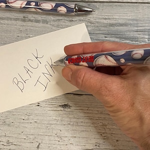 May include: A retractable ballpoint pen featuring a blue background with a repeating baseball pattern and the red text BEST COACH printed on the barrel. The pen has a silver-toned metal tip and clip, and it is shown writing on a white card with the text BLACK INK.