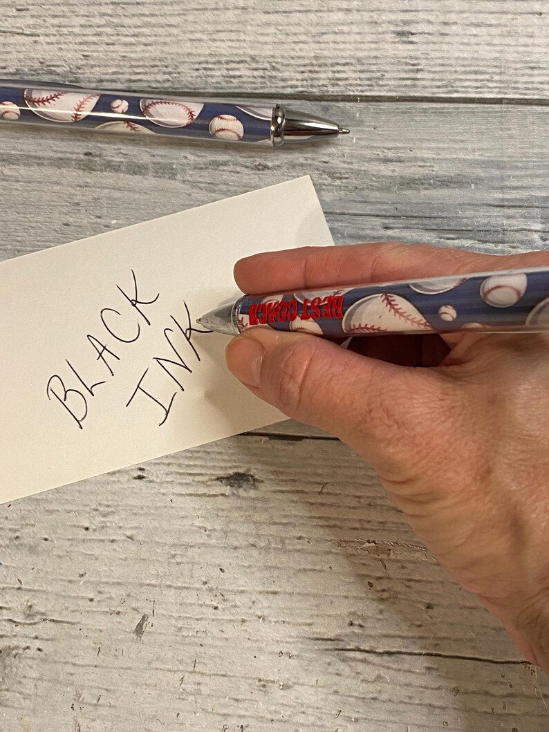 May include: A retractable ballpoint pen featuring a blue background with a repeating baseball pattern and the red text BEST COACH printed on the barrel. The pen has a silver-toned metal tip and clip, and it is shown writing on a white card with the text BLACK INK.