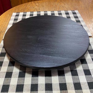 May include: A round, black wooden serving tray with a slightly raised edge. The tray is set on a black and white checkered placemat, creating a contrast in textures and patterns. The wood grain is visible on the tray's surface.