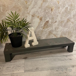 May include: A dark gray wooden riser with a rectangular top and two short legs. A potted green plant and a white wooden letter "A" are on the riser. The background is a marble-patterned wall.