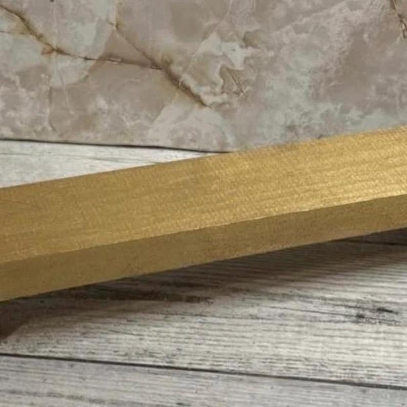 K&ouml;nnte beinhalten: Ein rechteckiger Holzblock mit einer glatten, metallisch goldenen Oberfl&auml;che. Der Block ist schr&auml;g positioniert und zeigt seine lange, flache Oberseite sowie die geraden Kanten, wobei das Licht auf der gleichm&auml;&szlig;igen, schimmernden goldenen Fl&auml;che reflektiert wird.