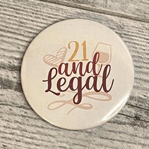 May include: A round button with the text "21 and Legal" in a cursive font. The number "21" is in gold, and the words "and Legal" are in a dark red color. The button also features a heart and a wine glass.