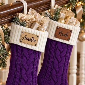 Puede incluir: Dos calcetines navideños de punto trenzado en color morado con puños acanalados blancos. Cada calcetín tiene una etiqueta rectangular de cuero sintético sujeta con cuatro remaches metálicos, con los nombres Amelia y Noah en letra negra.