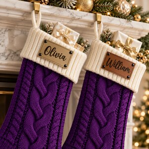 Puede incluir: Dos calcetines navideños de punto trenzado morado con puños acanalados de color crema. Cada calcetín tiene una etiqueta rectangular sujeta con cuatro remaches negros. Una etiqueta es dorada con el nombre Olivia y la otra es marrón con William.