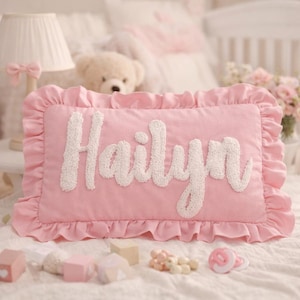 May include: A rectangular pink cushion featuring a ruffled border and the name Hailyn written in large, white, textured, raised script lettering across the centre. The cushion is made of a soft fabric and has a distinct, decorative, frilled edge.