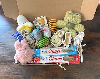 Puede incluir: Una caja de regalo llena de juguetes y golosinas para gatos. Incluye un peluche de rana, un plátano y un juguete rosa en forma de estrella. También presenta pegatinas con temática de gatos, muelles y dos paquetes de golosinas para gatos Inaba Churu.