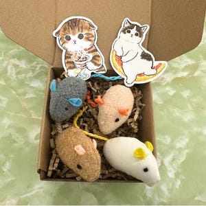 May include: A set of four plush cat toys in a cardboard box. The toys are shaped like mice and feature different colours and textures. The box is filled with brown packing paper.