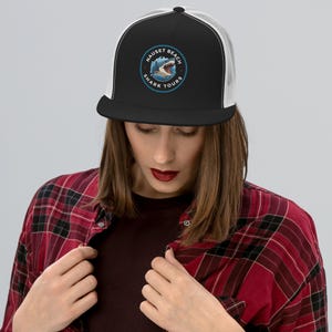May include: Black and white trucker hat with a circular logo featuring a shark and the text "NAUSET BEACH SHARK TOURS". The hat has a black brim and a white mesh back. The person is wearing a red and black plaid shirt.