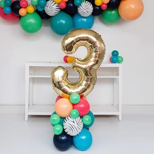 May include: A large gold foil balloon shaped like the number three, mounted on a vertical column of multi-colored balloons including shades of teal, pink, orange, dark blue, and white balloons with black zebra stripes, with a balloon arch above.