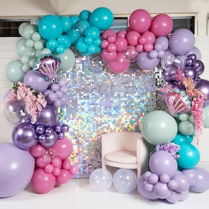 May include: A backdrop of iridescent sequins is decorated with a balloon arch in shades of purple, pink, teal, and green. The arch is made of various sizes and textures of balloons, including some with a metallic finish. A white chair sits in front of the backdrop.