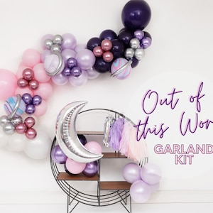 May include: A silver crescent moon balloon, pink, purple, and white balloons, and a silver and purple planet balloon are arranged in a garland. The garland is hanging on a white wall. The text "Out of this World Garland Kit" is written in purple on the wall.