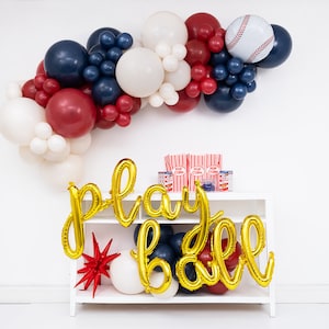 May include: A balloon arch in navy blue, burgundy, and white with a baseball-themed foil balloon. Gold foil script balloons spell out "play ball" in front of a white shelf containing red and white striped popcorn containers and small blue items.