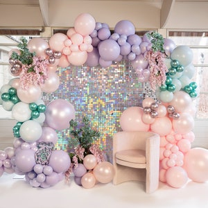 May include: A decorative balloon arch in pastel colors, including pink, purple, and green, with a sequined backdrop and a small chair. The arch is adorned with floral accents and silver orbs, creating a festive and elegant display.