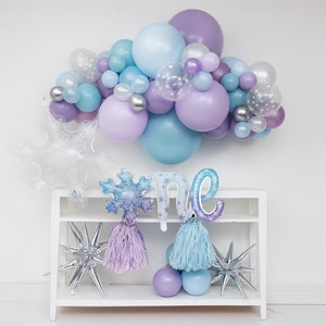 Puede incluir: Una estanter&iacute;a blanca con dos estantes contiene una guirnalda de globos azules y morados, un globo con forma de copo de nieve, un globo "uno" y dos globos con forma de estrella. Los globos tienen un tema invernal.