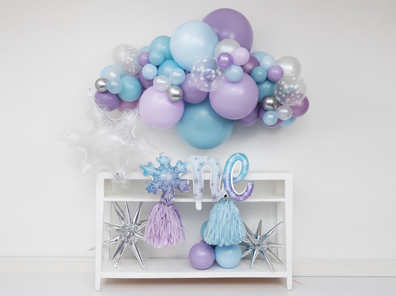 Puede incluir: Una estanter&iacute;a blanca con dos estantes contiene una guirnalda de globos azules y morados, un globo con forma de copo de nieve, un globo "uno" y dos globos con forma de estrella. Los globos tienen un tema invernal.
