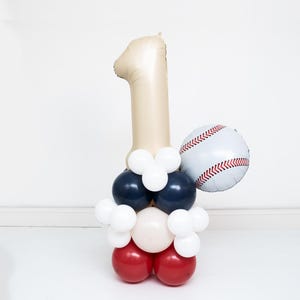 May include: A balloon sculpture featuring a large beige number one balloon at the top, a round white baseball balloon with red stitching on the side, and a base constructed from stacked red, navy blue, white, and beige round balloons.