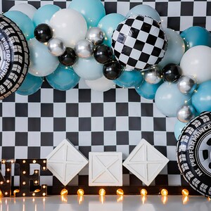 Pu&ograve; includere: Uno sfondo a scacchi bianco e nero con un arco di palloncini blu e bianchi. L'arco include palloncini a forma di pneumatici e una bandiera a scacchi. Il numero "UNO" &egrave; scritto in nero con luci bianche. Ci sono tre scatole bianche in primo piano.