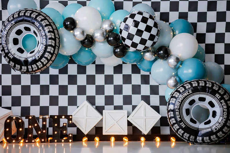 Pu&ograve; includere: Uno sfondo a scacchi bianco e nero con un arco di palloncini blu e bianchi. L'arco include palloncini a forma di pneumatici e una bandiera a scacchi. Il numero "UNO" &egrave; scritto in nero con luci bianche. Ci sono tre scatole bianche in primo piano.