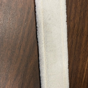 May include: Close-up of a white, rectangular, terry cloth headband. The headband is made of a soft, absorbent material and has a stitched border. The background is a dark brown wooden surface.
