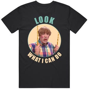 May include: Black t-shirt featuring a graphic of a person on a swing, with the text "LOOK WHAT I CAN DO" in a circular design. The person in the graphic is wearing a plaid shirt and has a surprised expression.