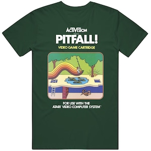 May include: Dark green t-shirt featuring the Activision logo and the text "PITFALL! VIDEO GAME CARTRIDGE." The graphic depicts a scene from the classic video game, with a character swinging over alligators. The shirt is designed for the Atari Video Computer System.