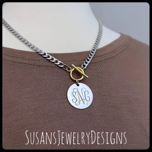 Op de afbeelding: Zilverkleurige gourmetketting met een goudkleurige toggle sluiting en een ronde hanger. De hanger heeft een gegraveerd monogram. De tekst "SUSANSJEWELRYDESIGNS" staat onderaan de afbeelding.