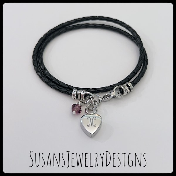 Pode incluir: Um bracelete trançado de couro preto com um fecho de coração prateado gravado com a letra "M". O fecho é preso a um fecho prateado e um pequeno cristal roxo.