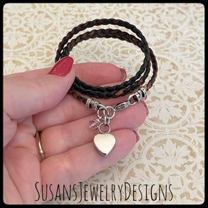 May include: A braided leather bracelet with a silver heart charm and a small crystal charm. The bracelet is brown and black and is shown on a hand.
