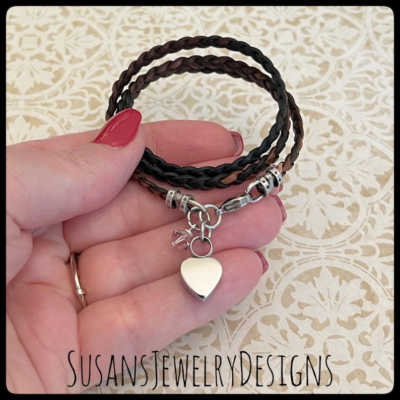 May include: A braided leather bracelet with a silver heart charm and a small crystal charm. The bracelet is brown and black and is shown on a hand.