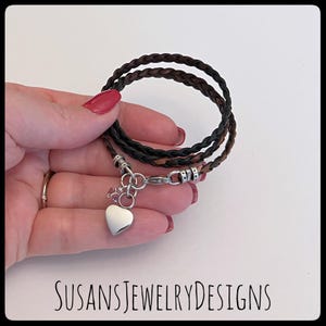 May include: A triple-strand leather bracelet with a braided design in brown and black. The bracelet has a silver heart charm with a small crystal accent.