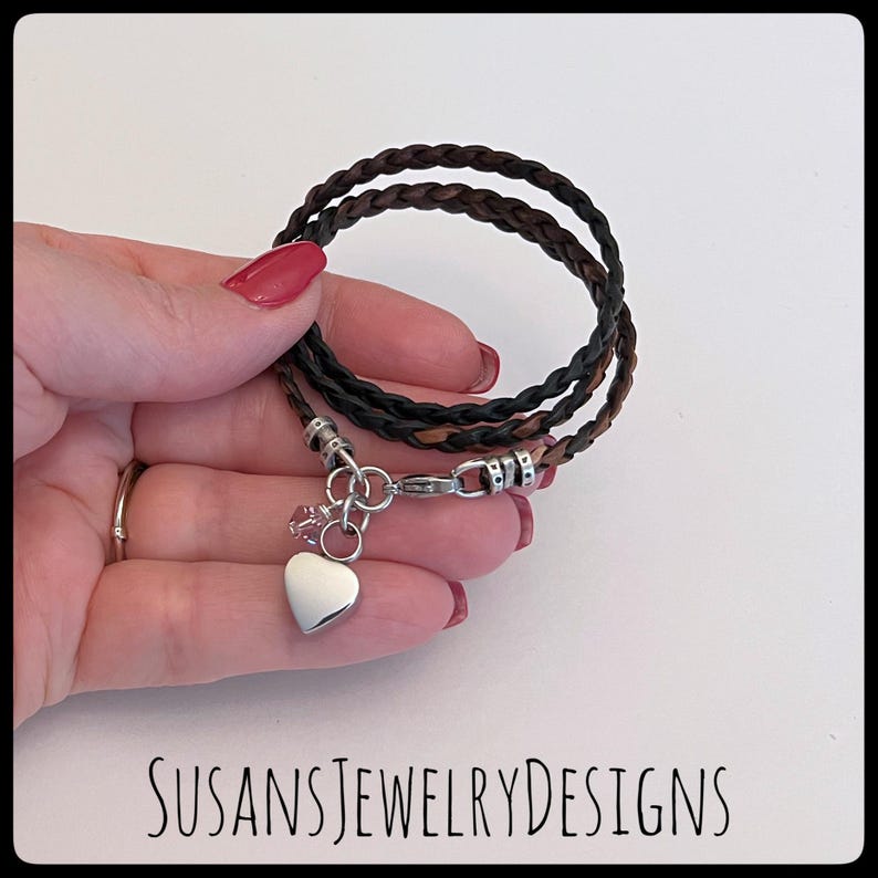 May include: A triple-strand leather bracelet with a braided design in brown and black. The bracelet has a silver heart charm with a small crystal accent.