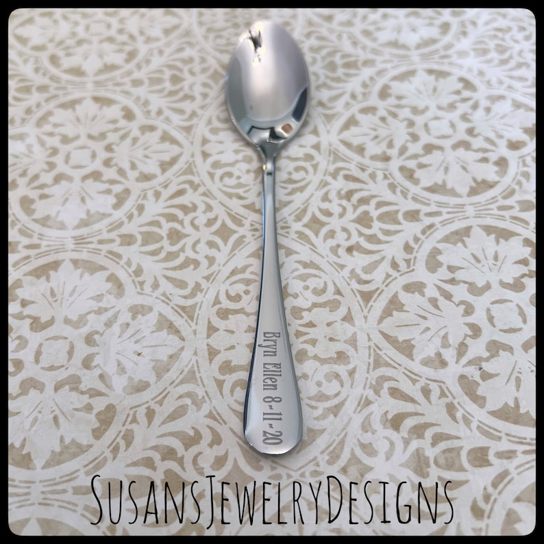 Puede incluir: Una cuchara plateada brillante con un mango largo. El mango est&aacute; grabado con el nombre "Bryn Ellen" y la fecha "8-11-20". La cuchara se muestra sobre una superficie crema estampada. El texto "SUSANSJEWELRYDESIGNS" est&aacute; en la parte inferior.