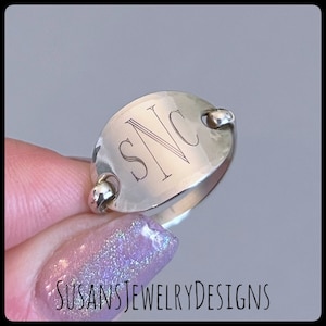 May include: A silver ring with an oval-shaped band and a hammered finish. The band is engraved with the initials "SNC".