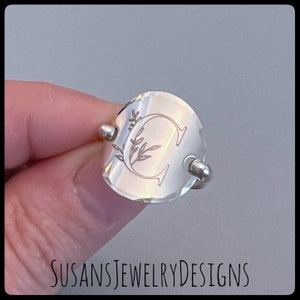 May include: A silver ring with a round, slightly heart-shaped face. The face is engraved with a stylized letter "C" and a floral design. The ring is on a finger.