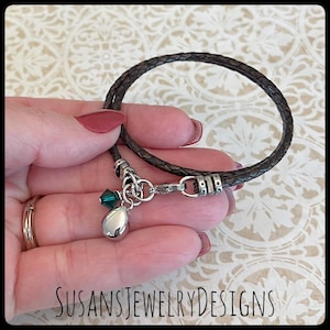 May include: A brown leather bracelet with a silver charm and green crystal accents. The bracelet has a silver clasp and is adjustable.