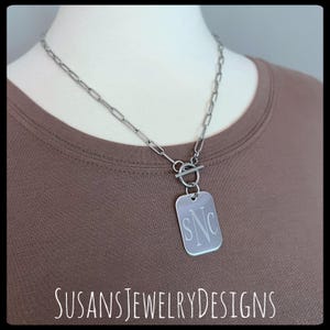 Op de afbeelding: Een zilveren ketting met een zilveren toggle-sluiting en een rechthoekige hanger gegraveerd met de initialen "SNC".