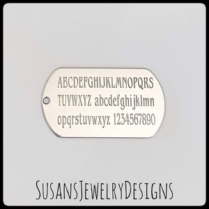 Puede incluir: Una placa de identificaci&oacute;n ovalada de color plateado con letras may&uacute;sculas y min&uacute;sculas grabadas, y n&uacute;meros. El texto "SUSANS JEWELRY DESIGNS" est&aacute; impreso en la parte inferior. La placa tiene un peque&ntilde;o agujero en el lado izquierdo.