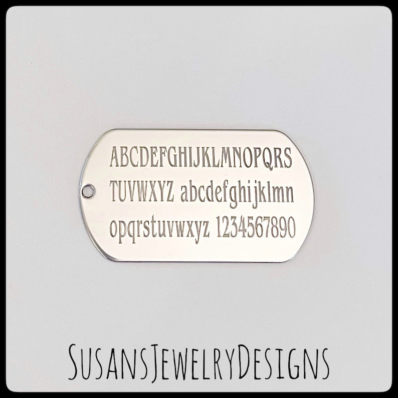 Puede incluir: Una placa de identificaci&oacute;n ovalada de color plateado con letras may&uacute;sculas y min&uacute;sculas grabadas, y n&uacute;meros. El texto "SUSANS JEWELRY DESIGNS" est&aacute; impreso en la parte inferior. La placa tiene un peque&ntilde;o agujero en el lado izquierdo.